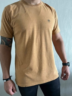 Homem vestindo uma camiseta mostarda com logo discreto, em um ambiente claro.