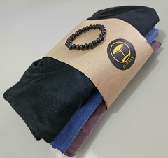 Conjunto de três camisetas empilhadas com uma pulseira preta, envolto em papel kraft com logotipo da marca Pinena.