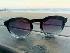 Óculos de sol unissex GRADE com armação preta e lentes escuras, em uma superfície de madeira, com o mar ao fundo.