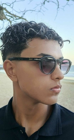 Jovem usando óculos de sol unissex GRADE na praia, com cabelo cacheado e camiseta escura.