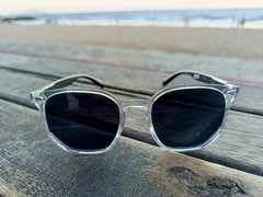 Óculos de sol hexagonais PARIS com armação transparente e lentes escuras, posicionados sobre uma mesa de madeira com fundo de praia.