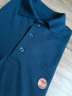 Camisa polo em tom de azul, com detalhes de botões e um emblema em couro.