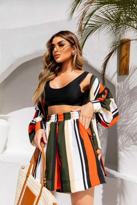 Mulher usando conjunto de camisa e shorts listrados em cores laranja, preto, verde e bege, com top preto e óculos de sol, segurando bolsa de palha