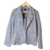 Blazer feminino de malha lilás acinzentado com bolsos frontais e lapela, pendurado em cabide de madeira