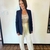 Mulher vestindo um blazer azul marinho com listras vermelhas nas mangas, acompanhado de uma blusa de tom neutro e calças claras, sorrindo em frente a uma parede clara.