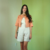 Mulher usando um conjunto de spencer salmão Jeane Steiner e shorts de linho claro, com colar colorido, em um fundo verde claro.