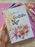Caderno da Gratidão com capa decorada por flores coloridas e uma borboleta, com fechamento espiral branco e pingente de tassel creme, sobre mesa com acessórios de papelaria