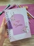 Caderno da Gratidão com capa decorada por flores e borboletas, segurado por uma mão, sobre uma mesa com outros materiais de papelaria.
