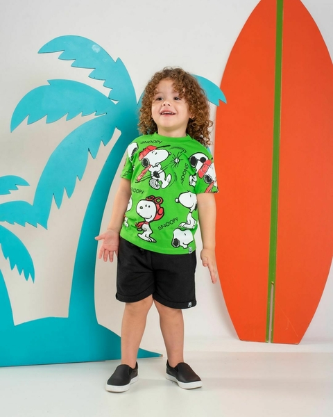 Criança sorridente vestindo uma camiseta verde com estampa do Snoopy, shorts pretos e tênis escuros, em um cenário com palmeiras e pranchas de surf coloridas.