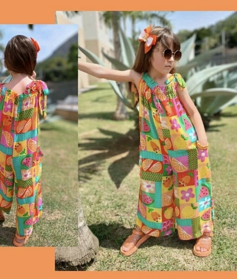 Menina vestindo um macacão colorido com estampas de frutas, posando ao ar livre com óculos de sol e um laço no cabelo.