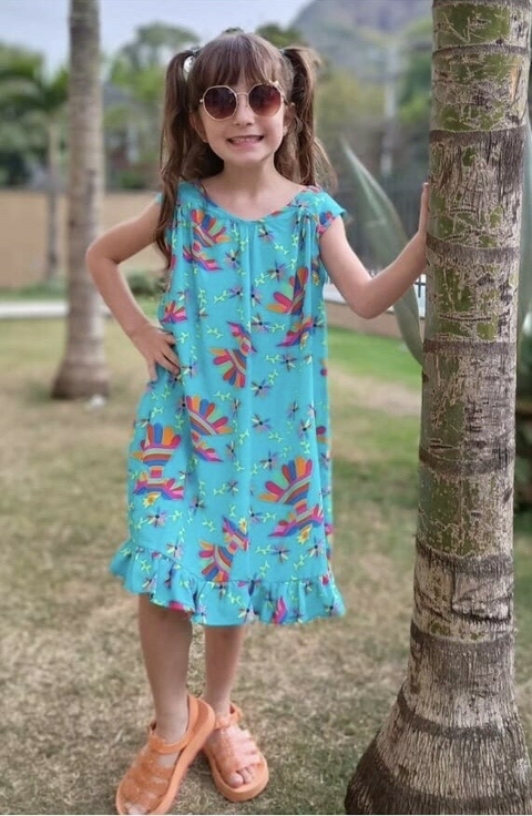 Menina sorridente usando um vestido azul com estampas coloridas, óculos de sol e chinelos, posando ao lado de uma árvore em um ambiente externo.