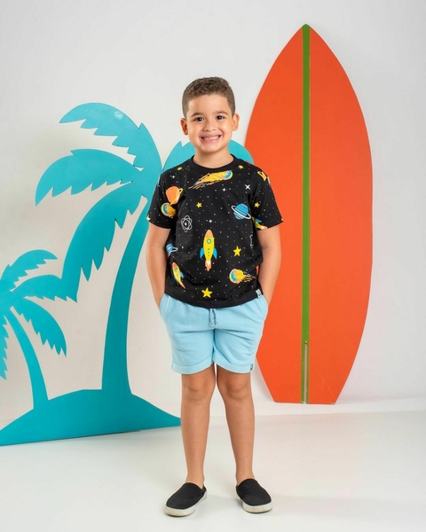 Criança sorridente usando camiseta preta com estampa de planetas e foguetes, ao lado de palmeiras e uma prancha de surf laranja.