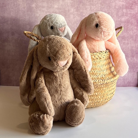 Tres conejitos de peluche en tonos beige, gris y rosa, sentados junto a una canasta de mimbre.