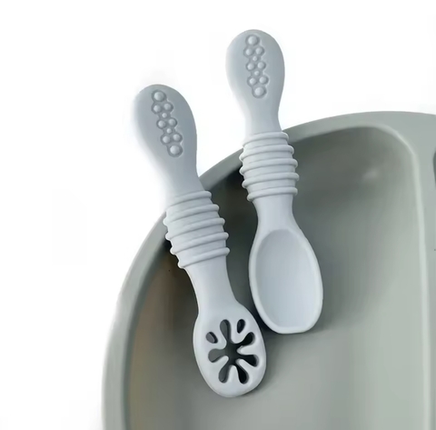 Juego de cubiertos de silicona en color blanco, con un diseño ergonómico y texturizado, colocados sobre un plato gris.