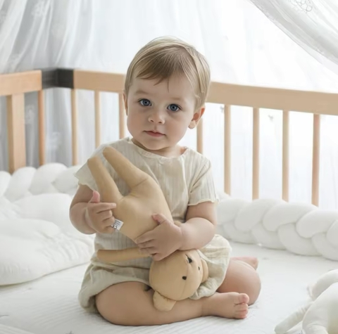 Bebé sentado en cuna blanca sosteniendo un muñeco de peluche color beige llamado Benito