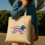 Tote bag beige con diseño divertido de perro salchicha estilo hot dog en colores azul y rosa, sostenido por una persona con camisa de jean