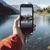 Uma mão segurando um smartphone com uma imagem de montanhas refletidas em um lago, enquanto um power bank solar está pendurado no dispositivo.