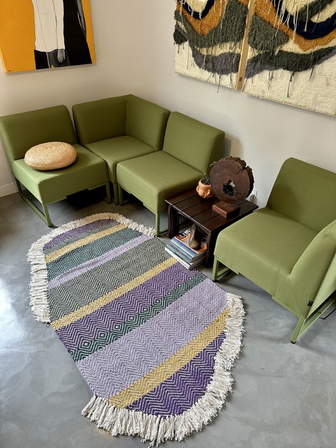 Tapeçaria Fauna em formato oval com franjas, apresentando padrões coloridos em um ambiente moderno.