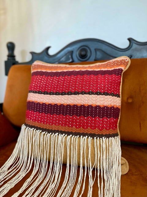 Almofada colorida com franjas, apresentando listras em tons de vermelho, rosa e marrom, repousando em um sofá marrom.
