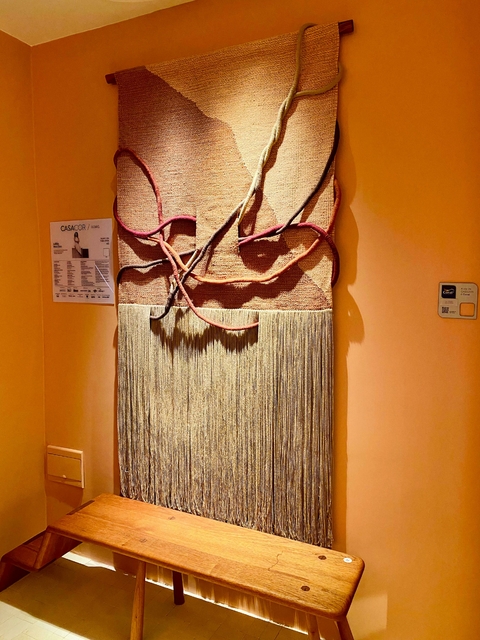 Arte têxtil chamada "Entre Tramas e o Tempo", destacando tramas e franjas, em uma parede de cor laranja, com um banco de madeira ao primeiro plano.