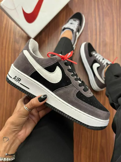 Tênis Nike Air Force 1 nas cores marrom e preto em camurça, sendo segurado por uma mão, com caixa de fundo.