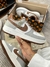 Tênis Louis Vuitton x Nike Air Force 1 nas cores cinza e marrom, segurados por uma mão. A caixa de fundo exibe o logotipo da Louis Vuitton.