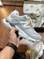 Tênis New Balance 9060 na cor cinza claro, sendo segurado por uma mão, com caixa ao fundo.