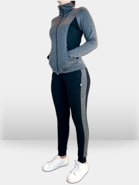 Foto de lado de uma mulher vestindo um agasalho em tecido Action Fleece Thermo, com jaqueta cinza mescla e detalhes em preto. A jaqueta possui gola alta, sem capuz, zíper frontal, bolsos laterais e recorte lateral preto. A calça é preta, com ribana na bar
