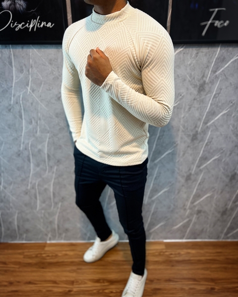 Homem usando um suéter raglan de tricô premium creme com elastano, exibindo um padrão em relevo e posando em um ambiente moderno.