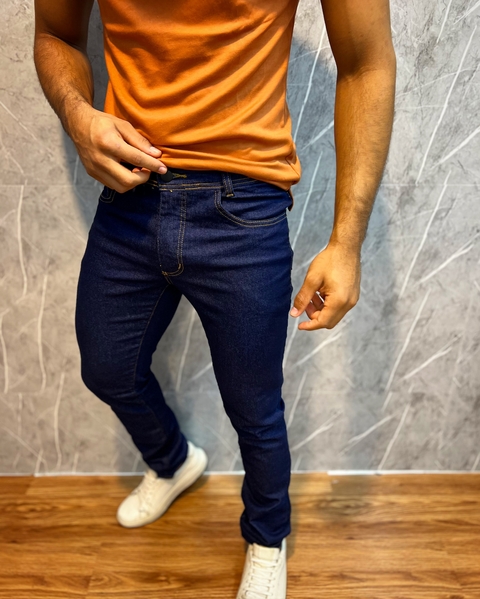 Homem vestindo calça jeans tradicional premium azul escura, camiseta laranja e tênis branco, em fundo cinza e chão de madeira