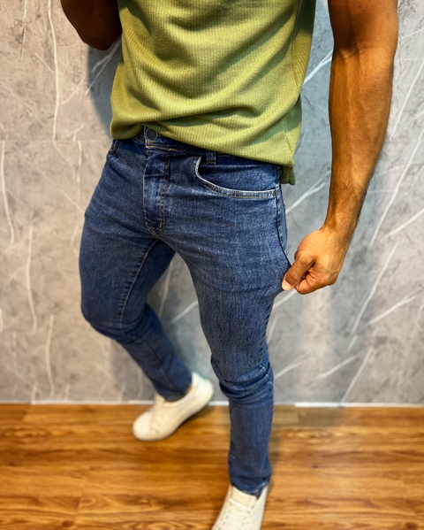Modelo usando calça jeans tradicional premium azul, com uma camiseta verde e tênis brancos, em um ambiente interno com parede texturizada.