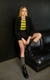 Modelo usando um blazer alfaiataria twill básico, camiseta listrada verde e preta, shorts pretos e botas brilhantes, sentada em um sofá preto.