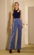 A imagem mostra uma modelo em pé, vestindo uma calça Street Cupro. A modelo combina a calça com uma blusa de tricot azul marinho com um recorte assimétrico no ombro, adornado com detalhes brilhantes. 
