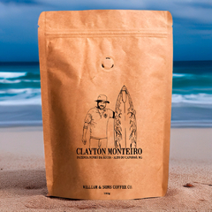 Café em grãos Clayton Monteiro 250g, da Fazenda Ninho da Águia, MG. Embalagem kraft ilustrada, fotografada com fundo de praia e mar azul vibrante.