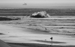 Surfer solitário caminha na praia em Chicama, Peru, com ondas alinhadas e embarcação ao fundo. Foto em preto e branco transmite poesia e imensidão.