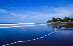 Praia de areia escura e mar azul com palmeiras ao fundo em um dia ensolarado. Refúgio tropical perfeito para relaxar ou surfar ondas consistentes. Km 59, em El Salvador