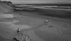 Praia deserta com longas ondas perfeitas e três surfistas caminhando com pranchas, capturada em preto e branco com atmosfera nostálgica e cinematográfica.