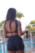 Mulher se alongando com um conjunto preto que inclui short fit, usando tênis branco, em ambiente externo com piscina ao fundo