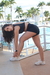 Mulher se alongando com um conjunto preto que inclui short fit, usando tênis branco, em ambiente externo com piscina ao fundo