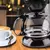 Detalle de la jarra de vidrio de la cafetera Dinax junto a una taza de café con espuma sobre mesa de madera.
