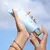 Imagem de uma mão segurando um tubo de hidratante Light Dream da coleção Brand Collection, com fundo de céu azul claro.