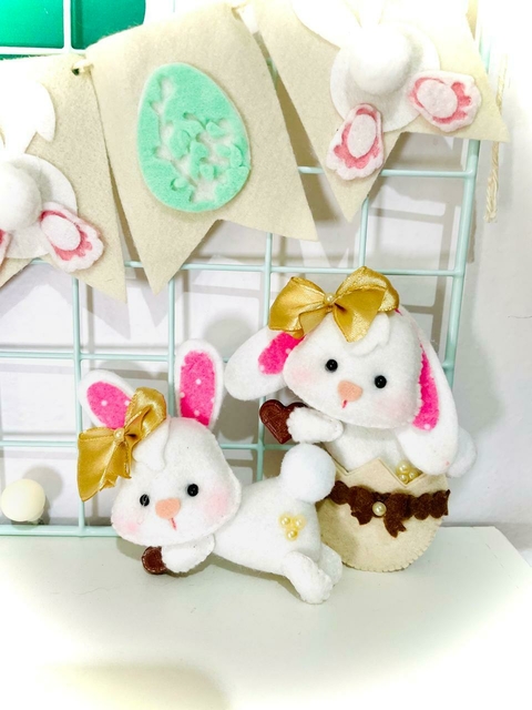 Coelhinhas de feltro decorativas segurando chocolates, com laços dourados e detalhes em rosa, perfeitas para a Páscoa.