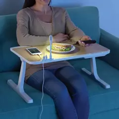 Mujer usando mesa multiusos para hogar con luz LED y cargador USB, mientras come un sándwich en el sofá
