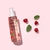 Frasco de fragancia corporal energizante con menta y frambuesa sobre fondo rosa, rodeado de hojas de menta y frambuesas frescas.