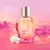 Perfume Autenti-k by me para mujer en botella facetada rosa con tapa plateada y flor blanca sobre fondo rosa