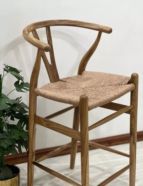 Banqueta Wishbone de madera con asiento tejido en ambiente interior junto a planta decorativa