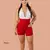 Mujer usando short tipo faja en neopreno de color rojo hasta media pierna, combinado con top deportivo blanca. Prenda diseñada para moldear el cuerpo, aplanar el abdomen y aumentar la sudoración durante el ejercicio.