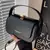 Bolsa feminina de ombro crossbody preta com alça única e detalhe dourado na tampa, sendo segurada por uma mão