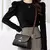 Mulher usando bolsa feminina de ombro em couro preto com detalhes marrons, combinando com blusa preta e saia xadrez