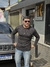 Homem usando suéter tricot preto, posando em frente a uma loja, com um carro Jeep ao fundo.